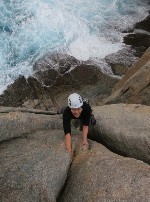 A woman climbing up a rock face with waves crashing under her